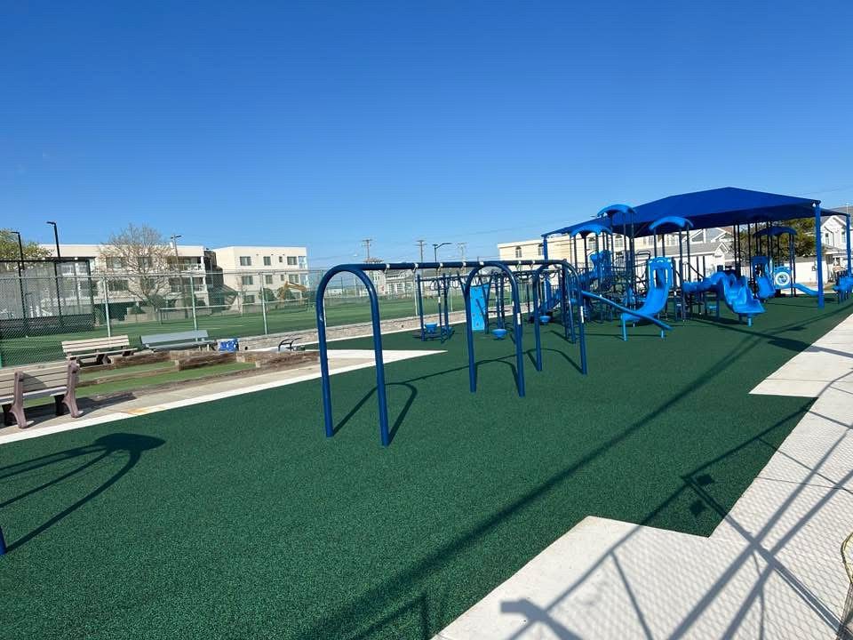 Blue playground equipment on green turf and a bocci court surrounded by cement paths and benches.
