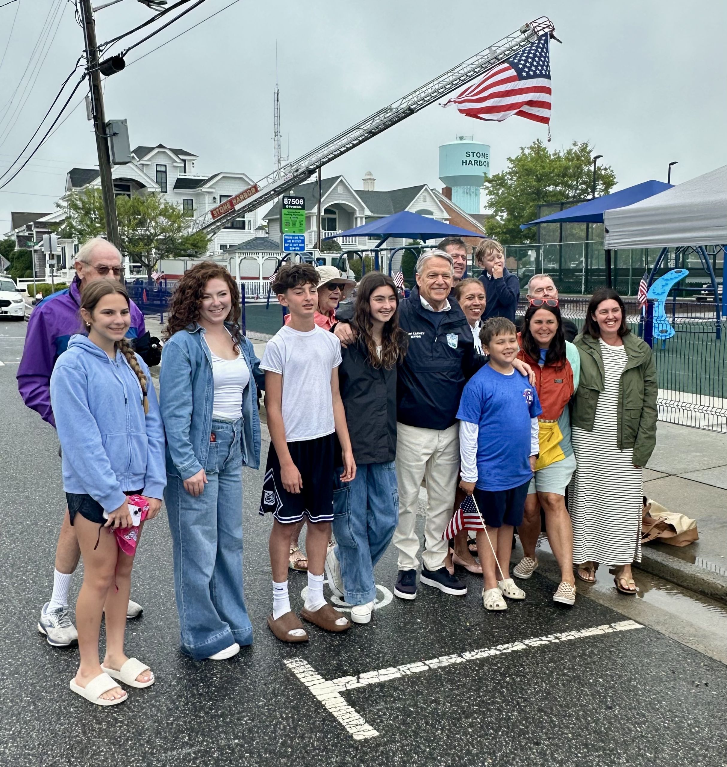 A cluster of smiling kids of many ages with the mayor and adults in front of flag on crane.
